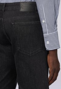 Vaqueros de denim negro con un acabado texturizado, que presentan un bolsillo trasero visible y una etiqueta de marca de cuero. La camiseta tiene rayas azules y blancas.