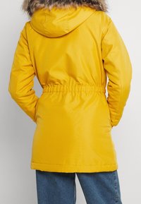 Parka amarilla con capucha forrada de piel, cintura elástica, exterior suave y mangas largas. Cuenta con un diseño de longitud media.