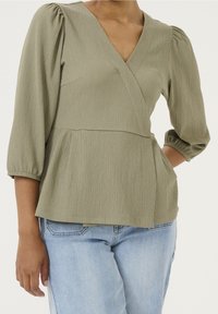 Olivgrüne, strukturierte Bluse mit V-Ausschnitt und Ballonärmeln, die eine taillierte Passform und eine A-Linien-Silhouette aufweist. Kombiniert mit hellblauen Jeans.