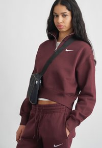 Sweatshirt scurt de culoare burgundy, cu guler înalt și logo-ul Nike argintiu. Asortat cu pantaloni de trening, la fel de burgundy, și o mică geantă crossbody neagră cu fermoare.