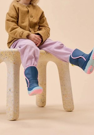 Niño con botas azules y rosas, chaqueta marrón y pantalones rosas sentado en un banco claro moteado con las piernas extendidas.