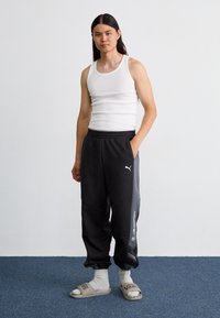 Pantalons de survêtement noirs avec logo PUMA, débardeur blanc et claquettes grises. Les pantalons de survêtement ont des poches et des poignets élastiques. Texture de tissu lisse et décontracté.