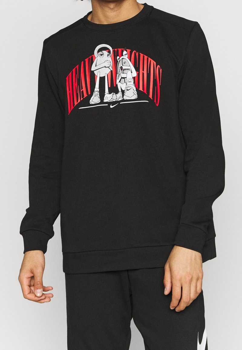 Personne portant un sweat-shirt noir avec le texte rouge "Heavy Heights" et des personnages de dessin animé au-dessus d'un petit logo blanc Nike sur la poitrine.