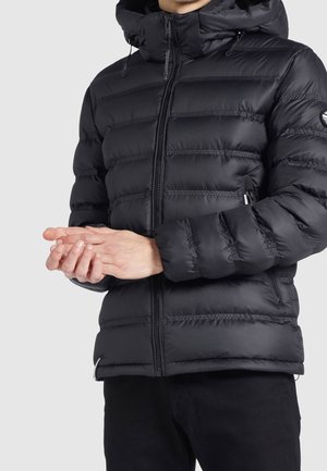 Czarna pikowana kurtka puchowa z przednim zamkiem błyskawicznym i kapturem, noszona przez osobę trzymającą ręce splecione na wysokości talii na szarym tle.