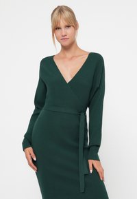 Robe en tricot à manches longues et enroulée, de couleur vert foncé, avec un décolleté en V et une taille nouée. Tissu texturé avec un motif côtelé. Silhouette ajustée.