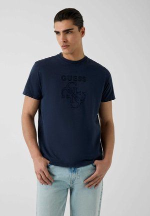 Jeune homme aux cheveux foncés portant un t-shirt bleu marine Guess et un jean bleu clair, debout avec les mains près des poches sur un fond uni.