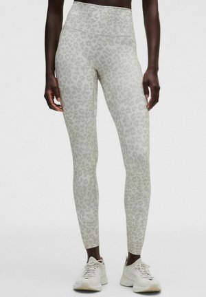 Leggings met hoge taille en lichtgrijze luipaardprint gedragen met witte sportschoenen door een donkerhuidig persoon die stil staat.