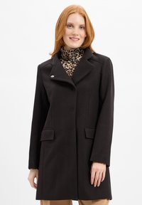 Zwarte wollen jas met een reverskraag, zijzakken en een decoratieve broche. Gekocht over een turtleneck met luipaardprint.