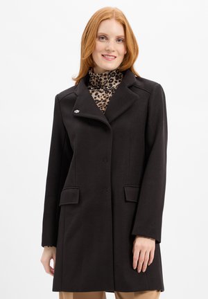 Schwarzer Wollmantel mit Reverskragen, Seitentaschen und einer dekorativen Brosche. Über einem Turtleneck mit Leopardenmuster getragen.