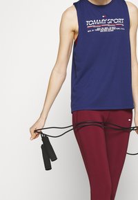 Top atletico senza maniche blu navy con il logo "TOMMY SPORT", abbinato a leggings bordeaux, mentre tiene una corda per saltare nera con impugnature testurizzate.