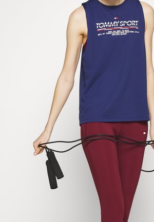 Marinblå ärmlös träningstopp med "TOMMY SPORT" logotyp, kombinerad med vinröda leggings, håller i ett svart hopprep med texturerade handtag.