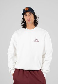 Giovane uomo con capelli lunghi che indossa un felpa bianca con un piccolo logo viola, pantaloni bordeaux e un cappellino nero con un emblema arancione su sfondo grigio.