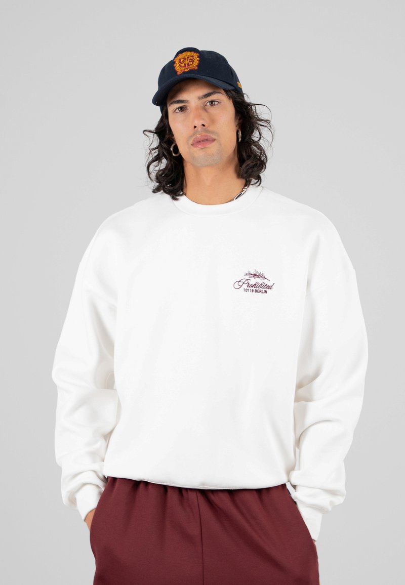 Giovane uomo con capelli lunghi che indossa un felpa bianca con un piccolo logo viola, pantaloni bordeaux e un cappellino nero con un emblema arancione su sfondo grigio.