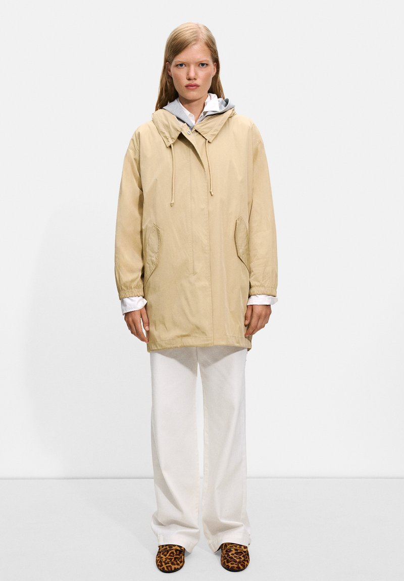 Veste beige légère avec capuche, poches avant et cordons de serrage, portée sur un sweat à capuche gris et un pantalon blanc, assortie à des chaussons à imprimé animal.