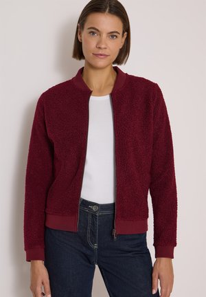 Femme portant une veste texturée bordeaux zippée sur une chemise blanche et un jean bleu foncé, debout devant un fond clair uni.