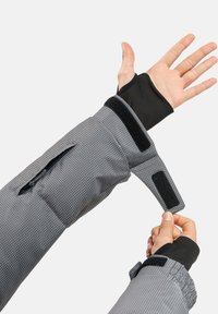 Manica grigia a motivo con accenti neri, dotata di tasca con zip e cinturino in Velcro, che presenta un materiale testurizzato e un design con foro per il pollice.