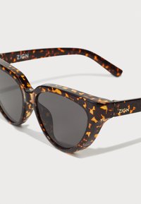 Tortoiseshell sunglasses with a bold, angular design. Black lenses, prominent golden logo on the temple, and textured frame accents.