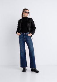 Μαύρο μπουφάν τύπου bomber, εφαρμοστή μαύρη ζιβάγκο, καμπάνα μπλε τζιν και μαύρα μποτάκια. Αξεσουάρ περιλαμβάνουν μεγάλα γυαλιά ηλίου και σκουλαρίκια.