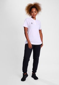 Witte polo met korte mouwen en een kraag, gecombineerd met zwarte trainingsbroeken en zwarte sneakers. Model staat recht vooruit.