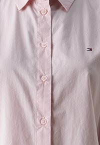 Lys pink skjorte med knapper, lavet af glat stof, med krave og et lille broderet logo i marineblå og rød på venstre side.