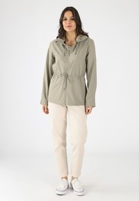 Chaqueta ligera con capucha en beige suave, con cintura ajustable con cordón y cierre frontal de cremallera, combinada con pantalones de color crema y zapatillas blancas.