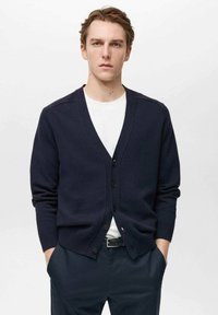 Mornarska jopica iz pletene tkanine z V-izrezom, zapiranjem na pet gumbov in dolgimi rokavi. Nosi se čez belo majico.