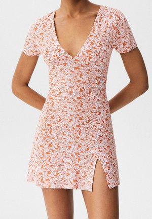 Femme portant une robe courte orange à fleurs avec un col en V, des manches courtes et une fente sur le devant, debout les mains dans le dos.