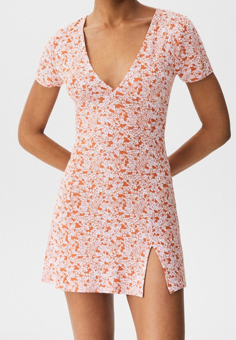 Femme portant une robe courte orange à fleurs avec un col en V, des manches courtes et une fente sur le devant, debout les mains dans le dos.