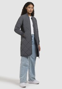Gequilted grijze lange jas met een ronde kraag, ritsluiting, zijzakken en geribbelde manchetten, gecombineerd met lichtblauwe wijde jeans en witte schoenen.