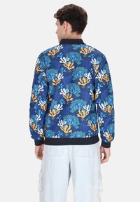 Heren bomberjack met een levendige print van een blauwe koraalrif, zwaar materiaal, geribbelde manchetten en broekrand, met oranje, teal en witte koraalaccenten.