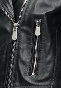 Κοντινό πλάνο μαύρου δερμάτινου μπουφάν με δύο κάθετα ασημένια φερμουάρ, ένα μεγαλύτερο κύριο φερμουάρ και ένα μικρότερο φερμουάρ τσέπης.