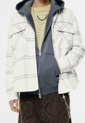 Person mit geschichteter Kleidung: grauer Zip-Hoodie, weiße karierte Hemdjacke, gestreiftes gelbes Shirt, Paisley-Hose und ein Sternketten-Accessoire.