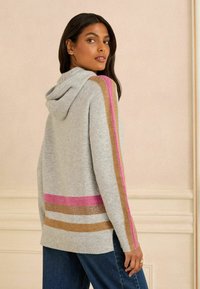 Szara bluza z kapturem z bocznymi paskami w kolorach różowym, złotym i białym. Miękka w dotyku, luźny krój, ściągacze na rękawach. Modelka stoi bokiem, widok od tyłu.