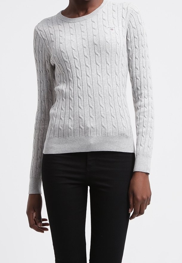 Femme portant un pull torsadé gris clair et un pantalon noir, debout devant un fond blanc uni.