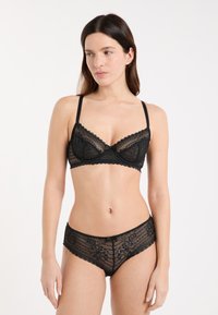 Woman wearing matching black lace bra and panties, standing against a plain white background, looking slightly left.