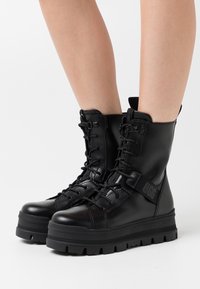 Bottes de combat en cuir noir avec fermeture à lacets sur le devant, semelle épaisse, et une sangle de marque. Présentent une finition lisse et des accents texturés.