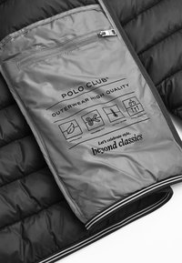 Sort quilted yderbeklædning med grå inderside; har en lynlås-lomme og trykte plejeanvisninger. Inkluderer branding og teksturerede detaljer.