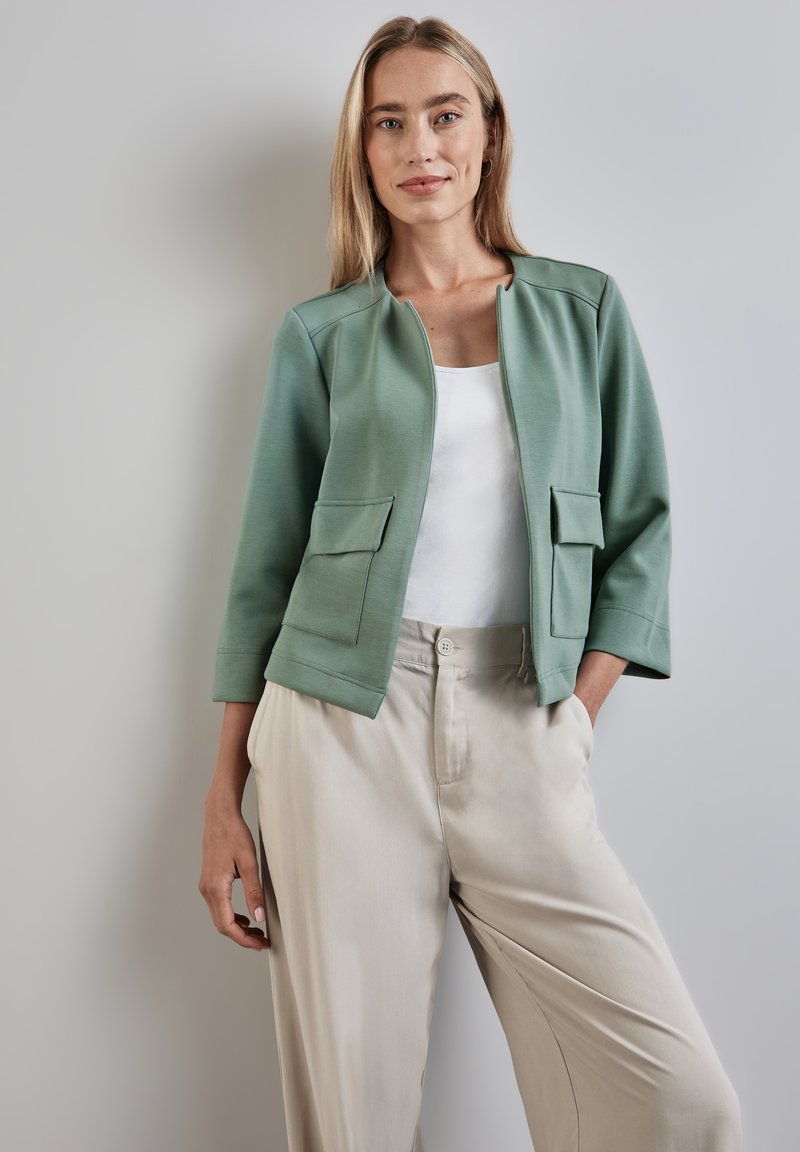 Veste structurée vert clair avec un col rond, des manches trois-quarts et des poches plaquées à l'avant, portée sur un haut blanc et un pantalon beige.