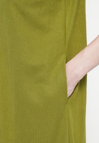 Oliven grønn kjole med sidelomme, laget av et mykt, teksturert stoff. Designet har en enkel, rettskåret silhuett.