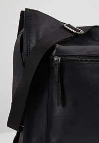 Sac en cuir noir avec une surface texturée, équipé d'une poche zippée et d'une bandoulière réglable avec une boucle en métal. Forme rectangulaire.