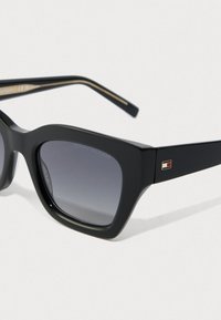 Sorte rektangulære solbriller med mørke gradientlinser og Tommy Hilfiger-logo på stængerne mod en ensfarvet hvid baggrund.