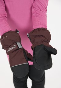 Moufles imperméables marron et noir avec une manchette, un poignet élastique et un détail de logo. Portées par une personne dans un haut à manches longues rose.
