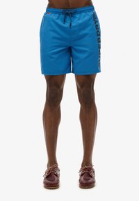 Shorts de bain bleus avec un cordon de serrage à la taille, présentant la marque "SUPERDRY" imprimée verticalement en noir sur le côté gauche.