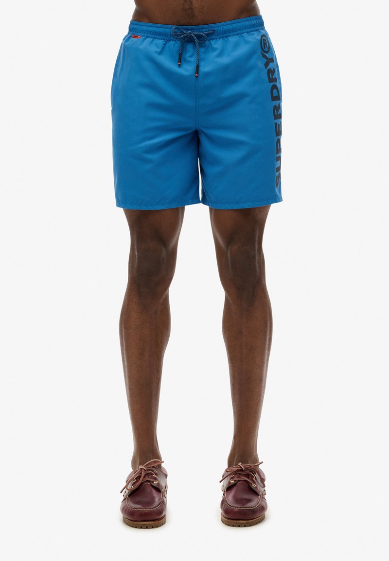 Shorts de bain bleus avec un cordon de serrage à la taille, présentant la marque "SUPERDRY" imprimée verticalement en noir sur le côté gauche.