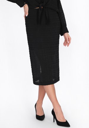 Frau trägt einen schwarzen, strukturierten Midirock, ein schwarzes Langarmoberteil, das an der Taille gebunden ist, und schwarze High-Heels, vor einem weißen Hintergrund.