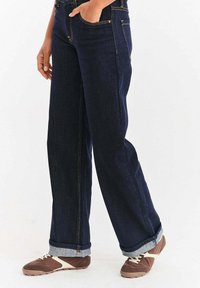 Osoba v tmavě modrých džínech s širokými nohavicemi zvednutými nahoře a hnědých a bílých sportovních teniskách, jedna ruka v kapse, na jednobarevném pozadí.