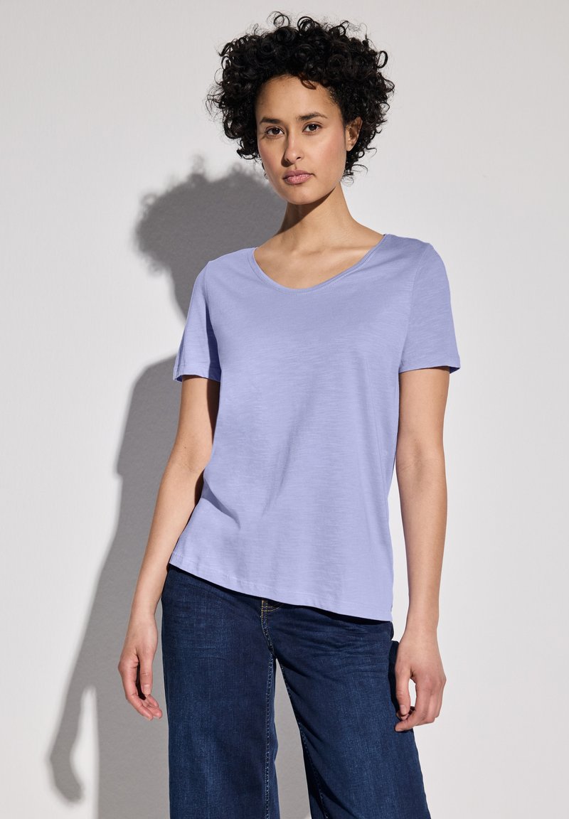 Camiseta de manga corta color púrpura claro con cuello redondeado, que presenta una textura suave y un corte relajado, combinada con jeans de mezclilla azul oscuro.