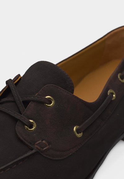 Σκούρο καφέ δερμάτινο παπούτσι σκάφους με ραφή μοκασίνου, δερμάτινα κορδόνια περασμένα μέσα από ορειχάλκινα μεταλλικά μάτια και ταμπά εσωτερική επένδυση.