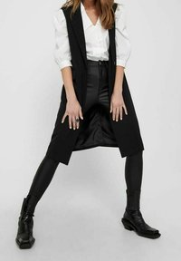 Czarna kamizelka bez rękawów z otwartym przodem, warstwowa na białej koszuli i czarnych skórzanych spodniach, noszona z czarnymi botkami za kostkę. Teksturowana tkanina.