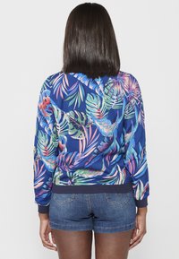 Giacca bomber blu navy con vivace stampa tropicale di foglie, polsini e orlo a costine. Tessuto leggero con una texture liscia.